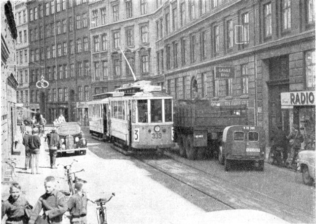 (Foto). Sporvognstog (toakslede vogne fra før første verdenskrig) på ensrettet sporstrækning i en af Nørrebros snævre gader (Elmegade) med parkerede biler.