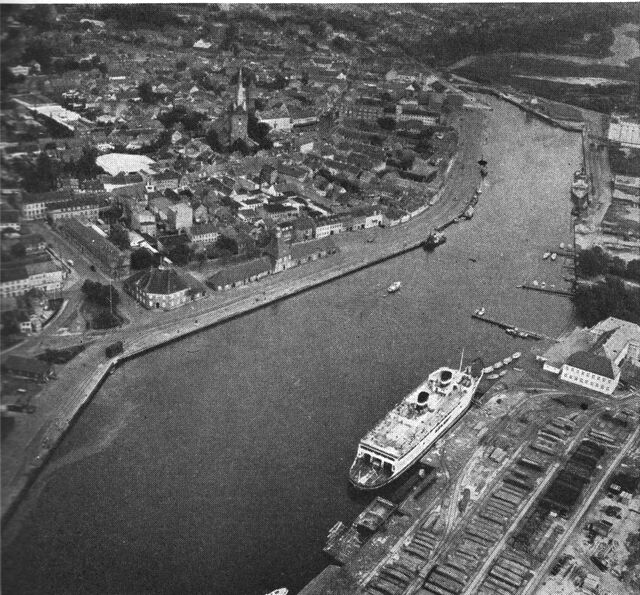 (Foto). Nakskov set fra luften fra vest. I forgrunden til højre et hjørne af skibsværftet (med en Storebæltsfærge til eftersyn ved kajen); på den anden side af havnen den gamle bykerne med kirketårnet i midten; øverst til højre ses i fortsættelse af havnen den bugtede Indrefjord.