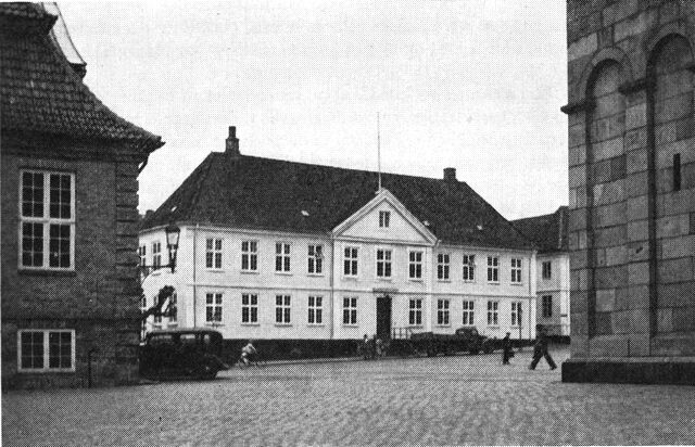 (Foto). Den gamle katedralskole, Skt. Mogensgade 1 (nu kommunal administrationsbygning) set fra sydøst over Gammeltorv. Til højre et hjørne af domkirken, til venstre museet, det gamle rådhus.