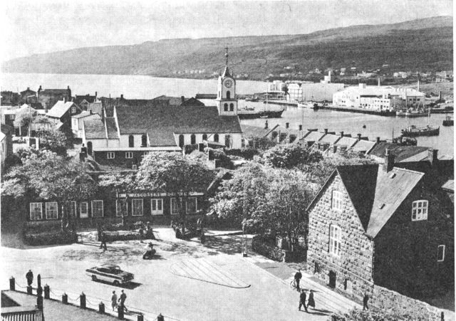 (Foto). Udsigt over en del af Tórshavn og Vestara Vág. Bag havneanlægget til højre fiskeriindustribygninger; i midten kirken; i forgrunden til højre den gamle kommuneskole fra 1894 og til venstre H. N. Jacobsens Bókahandil i den lange, lave tidligere realskolebygning fra 1860.